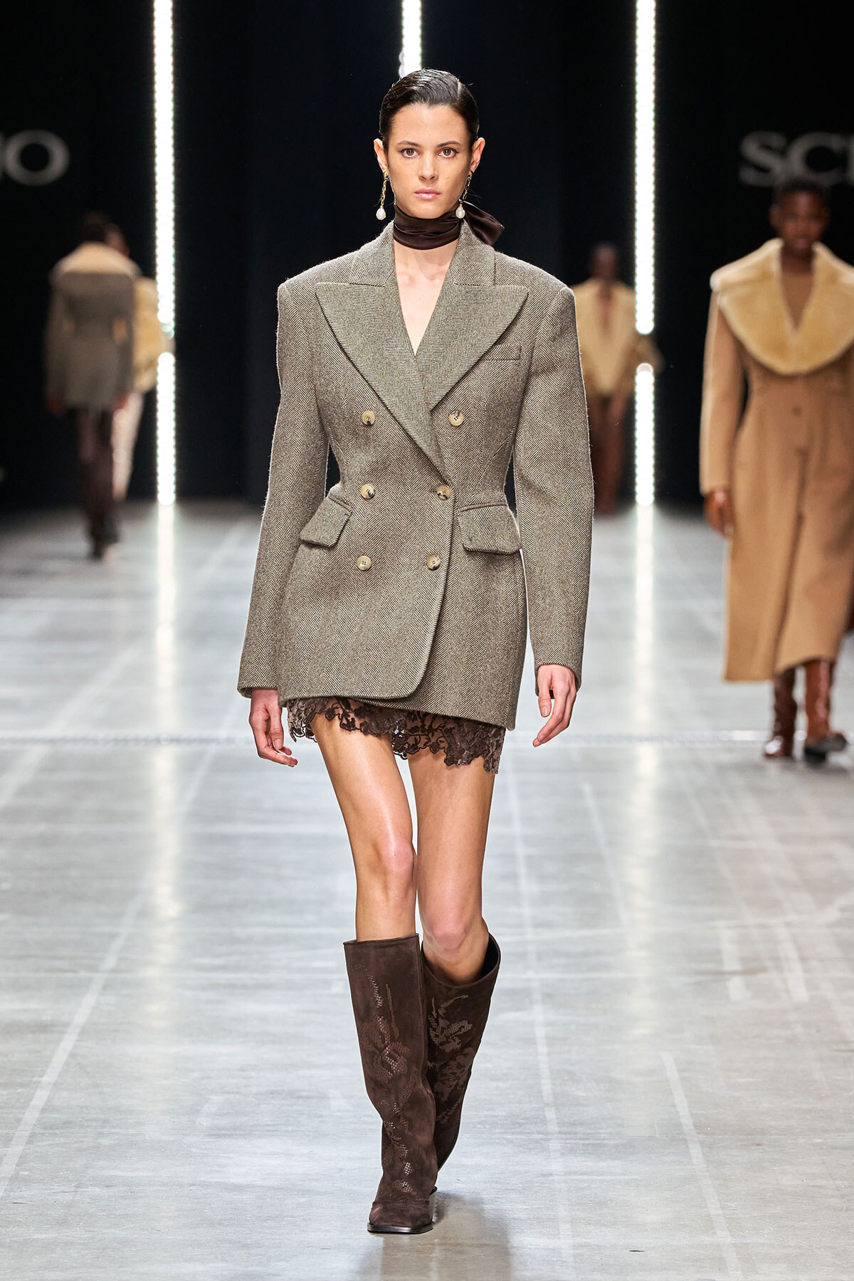 Model walking a runway in a gray tweed double-breasted blazer-dress, with a lace hem peeking below, paired with tall brown suede boots and a dark neck scarf.