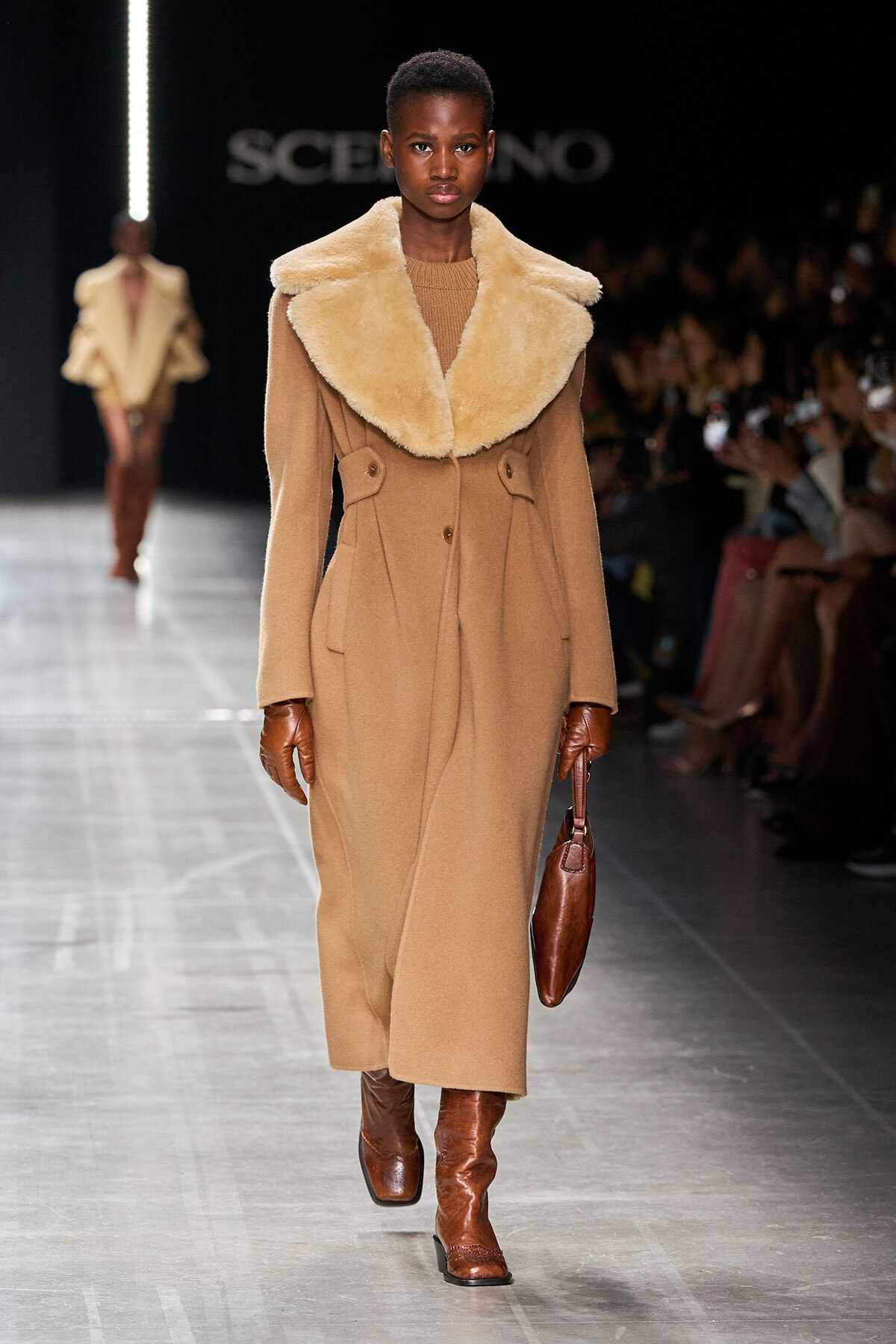 Full-length camel coat with a large cream shearling collar, brown gloves, and matching boots, carrying a brown handbag on the runway.