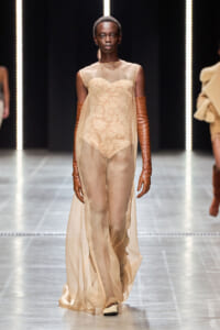 Model walking the runway in a sheer beige gown with a lace bodice and long brown leather gloves.