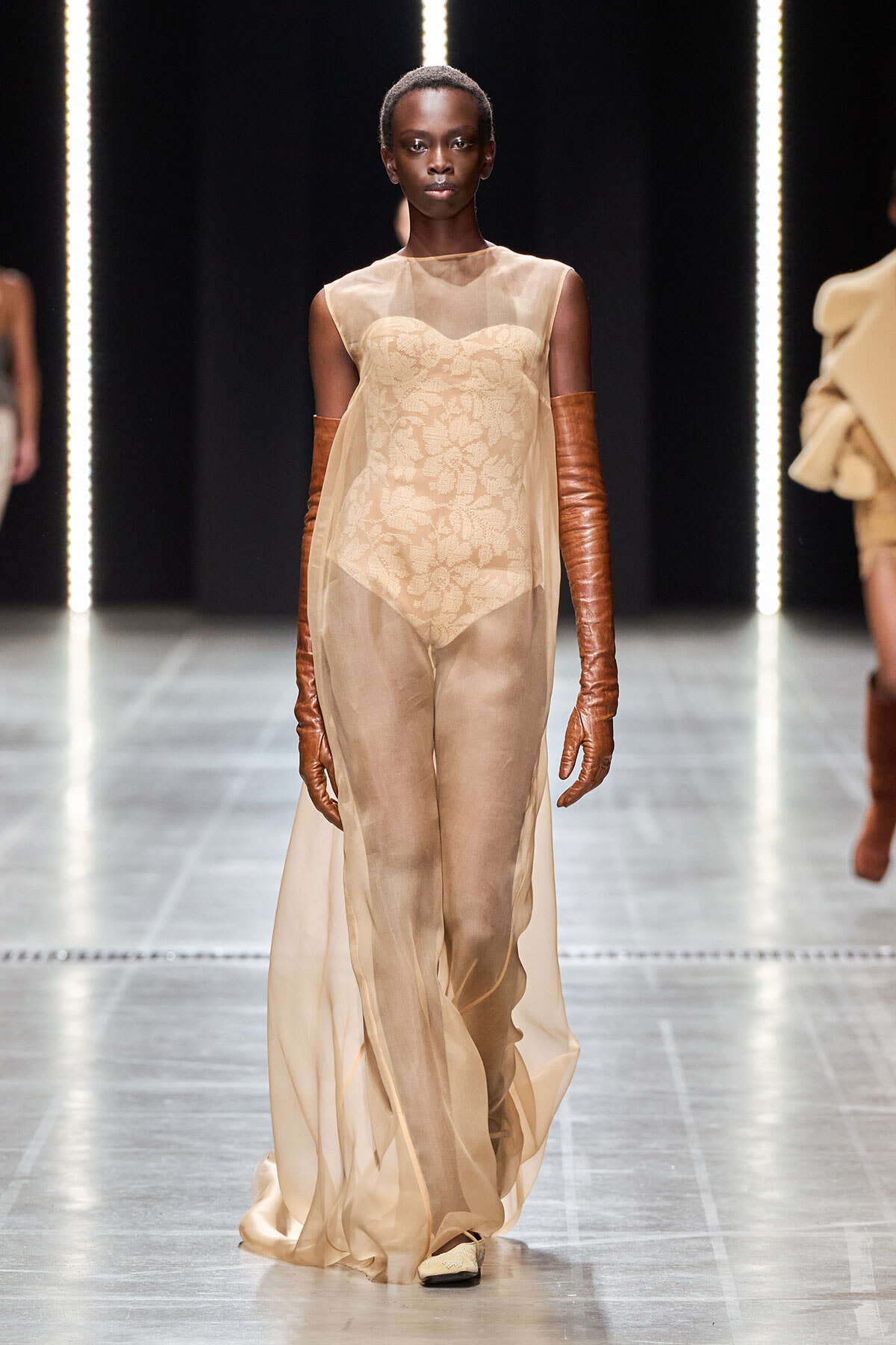 Model walking the runway in a sheer beige gown with a lace bodice and long brown leather gloves.