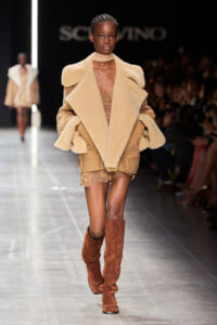 Model on a runway wearing an oversized beige shearling coat, lace-trim shorts, and tall brown suede boots.