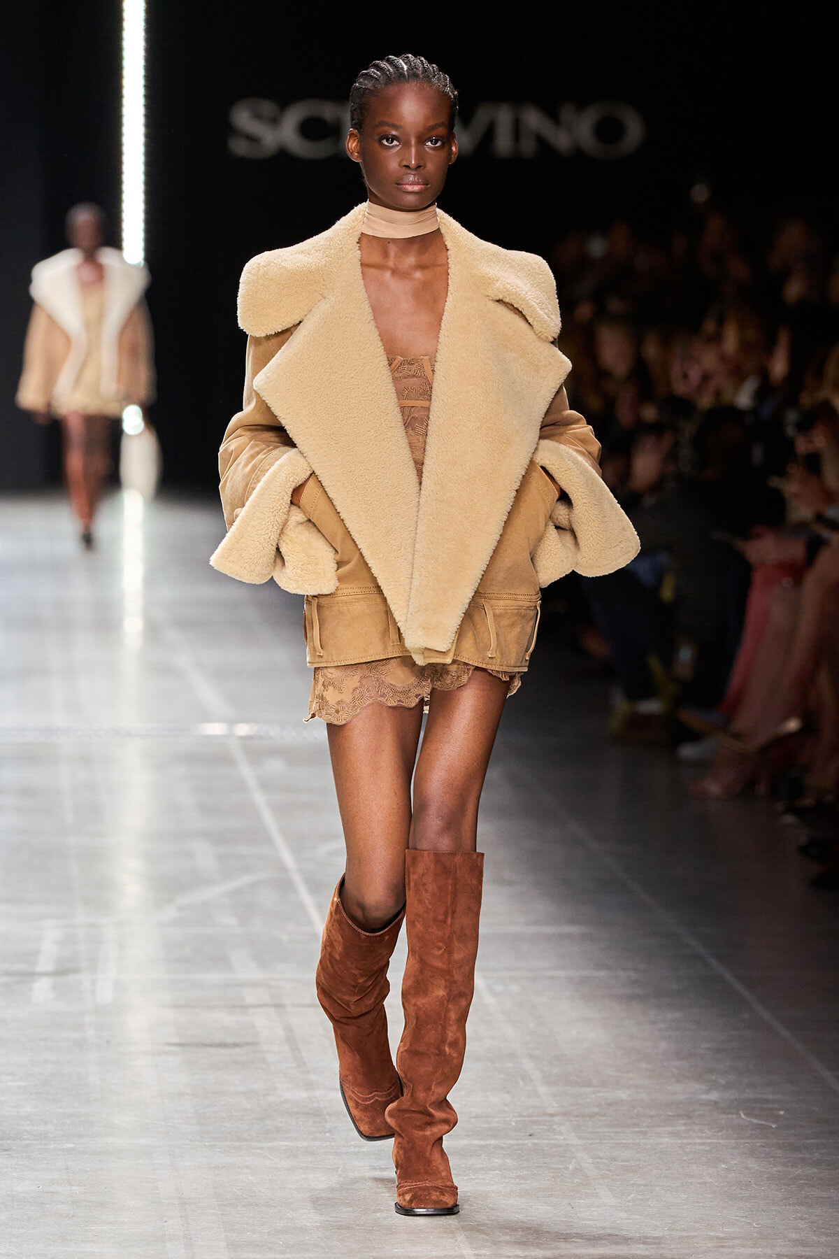 Model on a runway wearing an oversized beige shearling coat, lace-trim shorts, and tall brown suede boots.