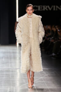 Model on a runway wearing a long cream boucle coat over a chunky knit sweater and skirt, with metallic sandals.
