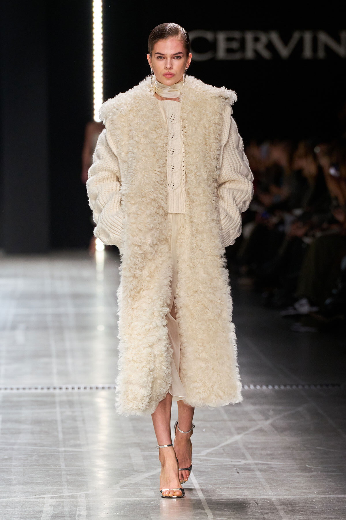 Model on a runway wearing a long cream boucle coat over a chunky knit sweater and skirt, with metallic sandals.