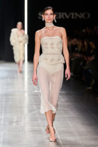 Runway model wearing a cream crochet strapless top with sheer ivory pants and silver sandals.