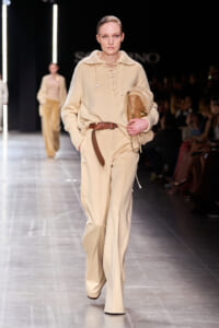Beige-toned fashion model on the runway wearing a knit sweater and cream wide-leg pants, carrying a tan fur-trimmed clutch.