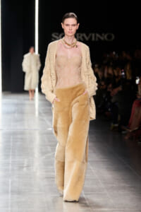 Runway model in nude-toned outfit: sheer bustier corset, chunky knit cardigan, and oversized tan faux-fur pants with a gold necklace.