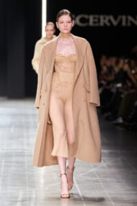 Model walking a fashion runway in a nude lace bodysuit with a long camel coat and strappy heels, neutral tones.