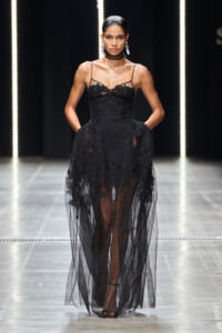 Model walking a fashion runway in a black lace gown with sheer tulle overlay, high waist, and thin straps, paired with dangling earrings and a choker, standing against a dark backdrop with vertical lights.