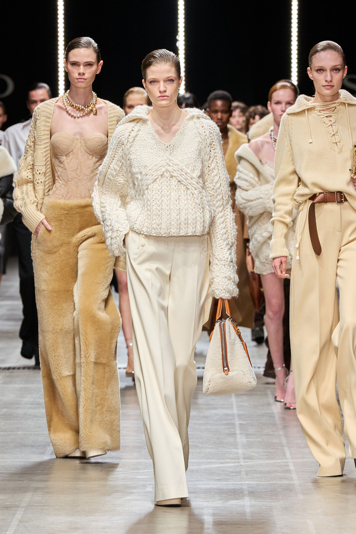Models walk a fashion runway in beige knitwear; center model wears a chunky cream sweater with wide trousers and carries a textured bag.