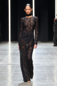 Model walking a runway in a full-length black lace gown with sheer sleeves and a choker neckline.