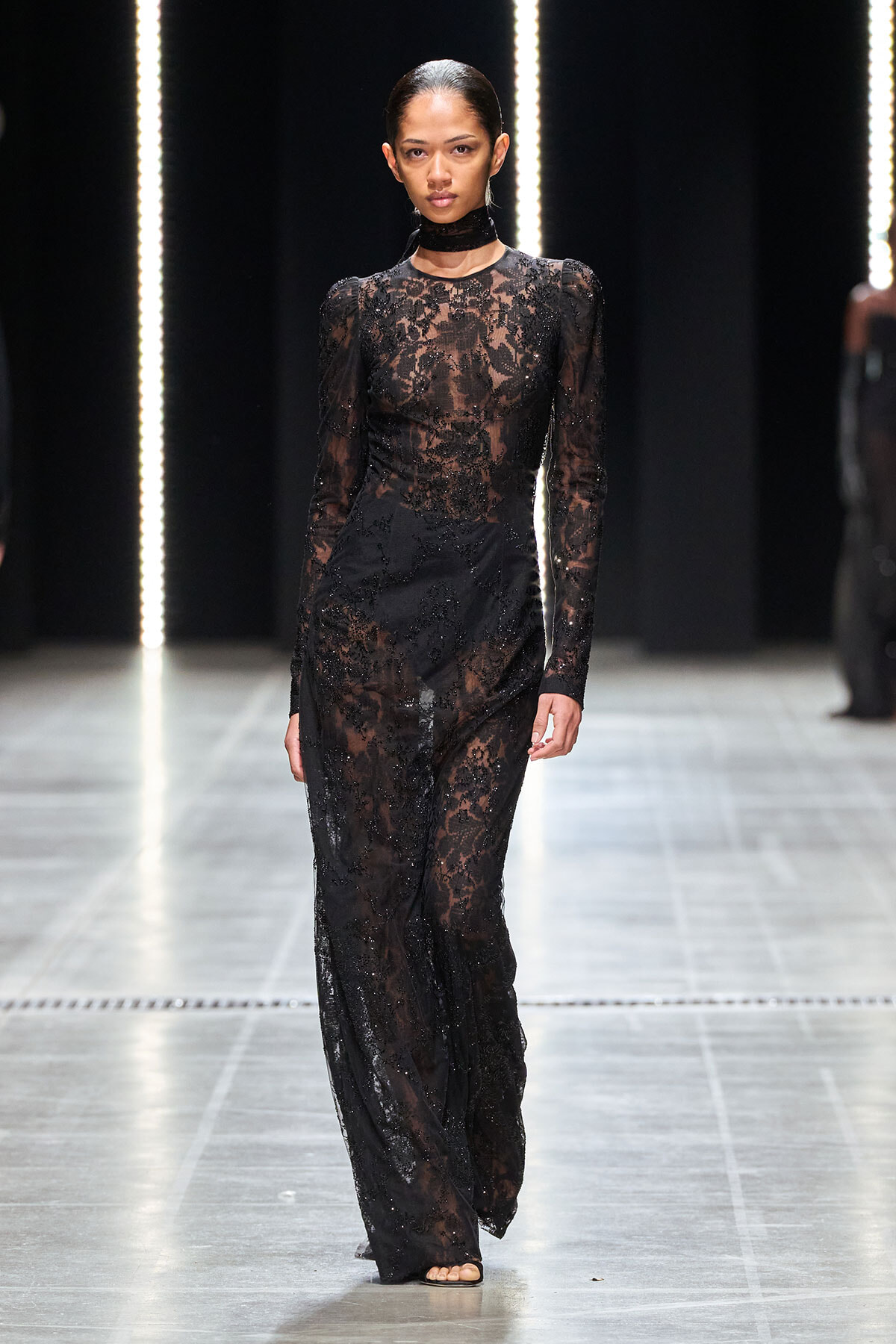 Model walking a runway in a full-length black lace gown with sheer sleeves and a choker neckline.
