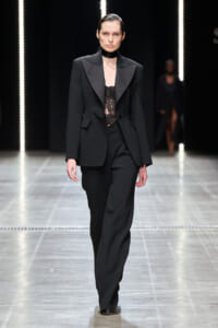 Model walks the runway in a black tailored suit with lace top and choker, under vertical light strips.
