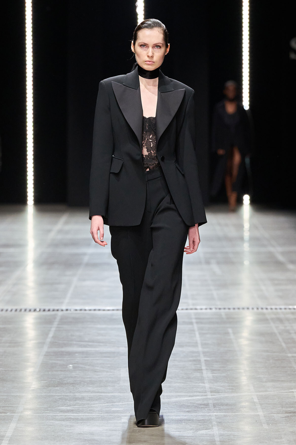 Model walks the runway in a black tailored suit with lace top and choker, under vertical light strips.