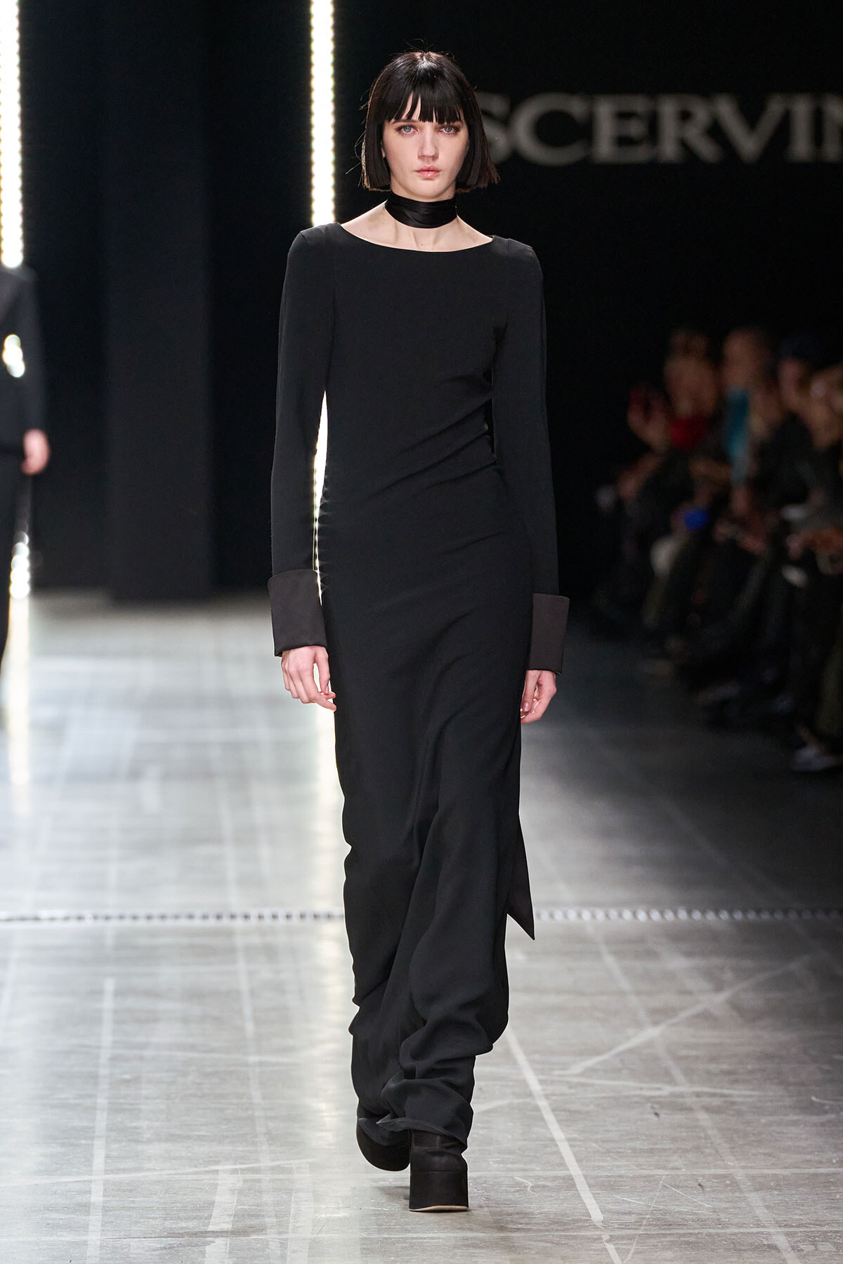 Model walking a runway in a long black dress with wide, cuffed sleeves and a black choker.