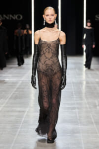 Model walks the runway in a sheer black lace gown with thin straps and long black gloves on both arms.