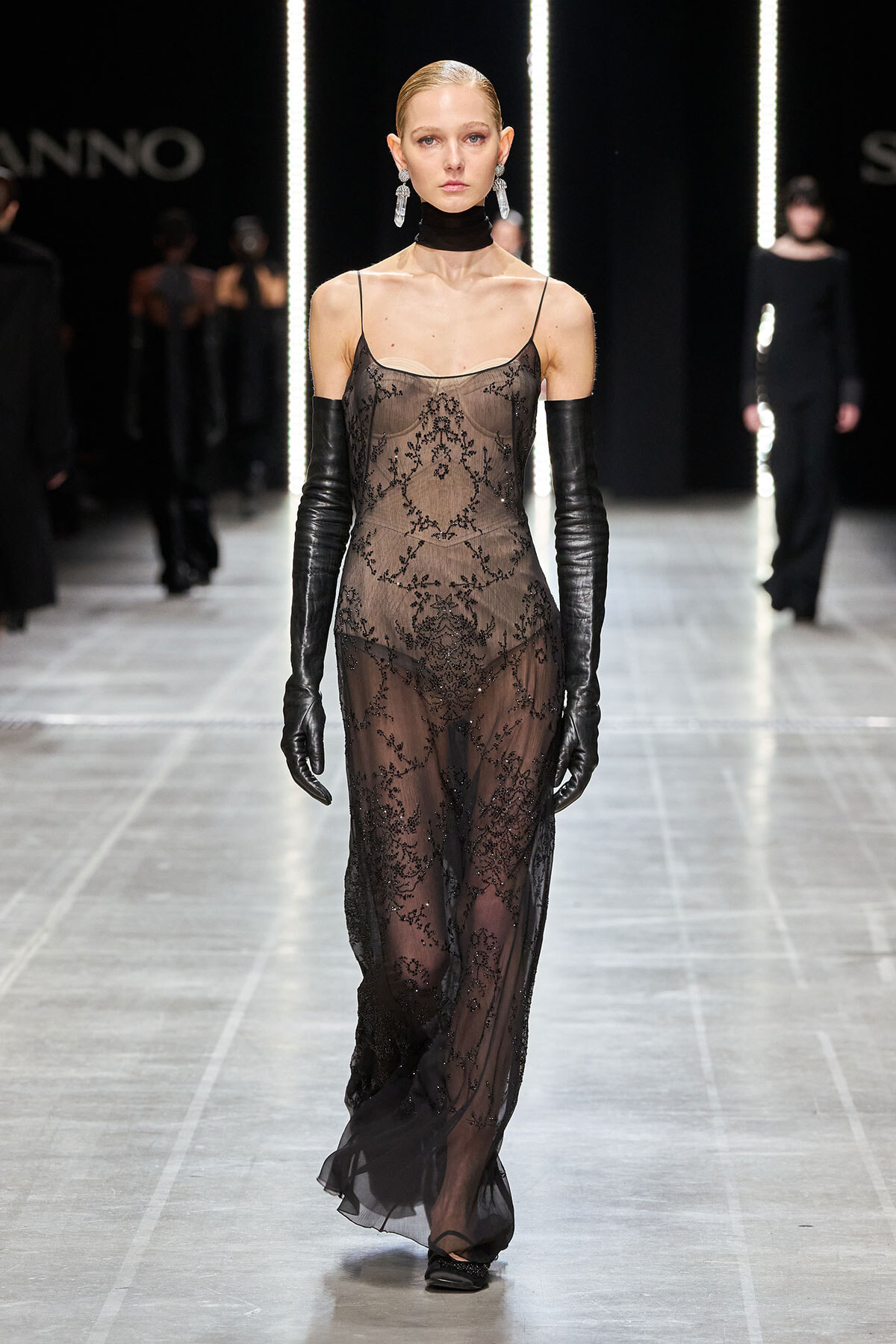 Model walks the runway in a sheer black lace gown with thin straps and long black gloves on both arms.