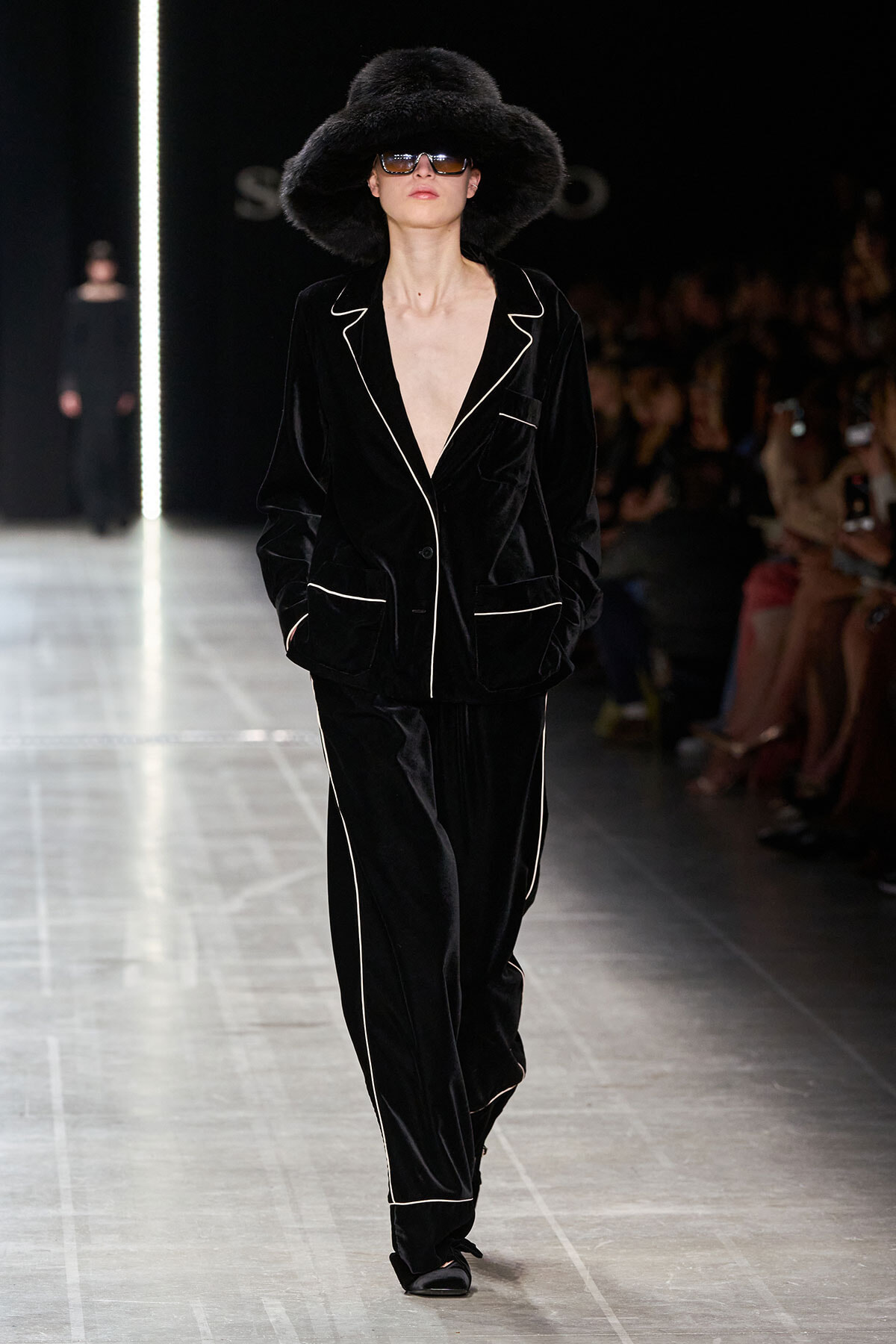 Model on a runway wearing a black velvet suit with white piping, oversized hat, and sunglasses.