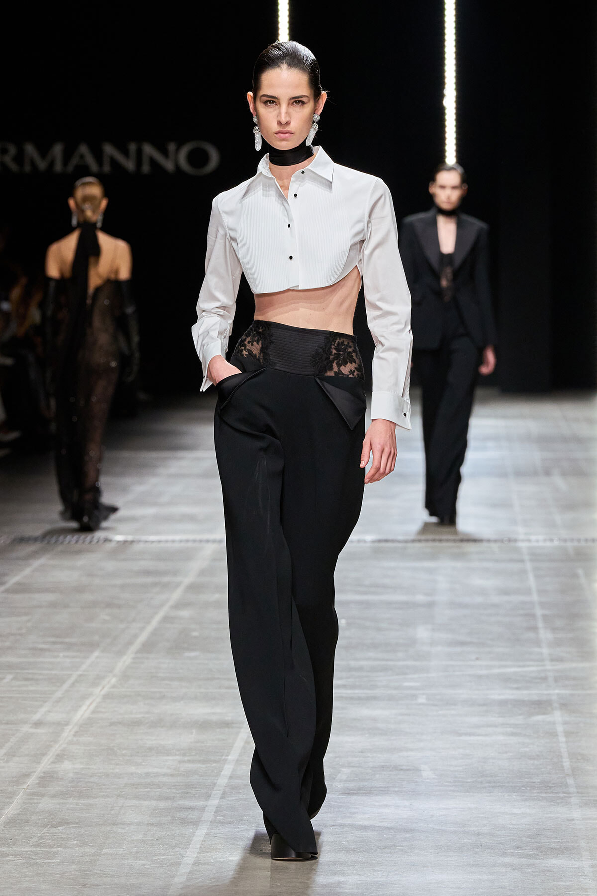 Model walks the runway in a white cropped shirt and black high-waisted pants with a lace waist panel.