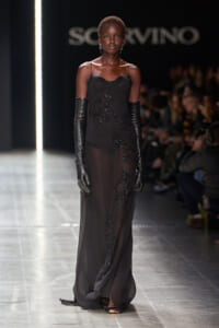 Model walks the runway in a black sheer evening gown with bead embroidery and long black gloves, against a fashion show backdrop.