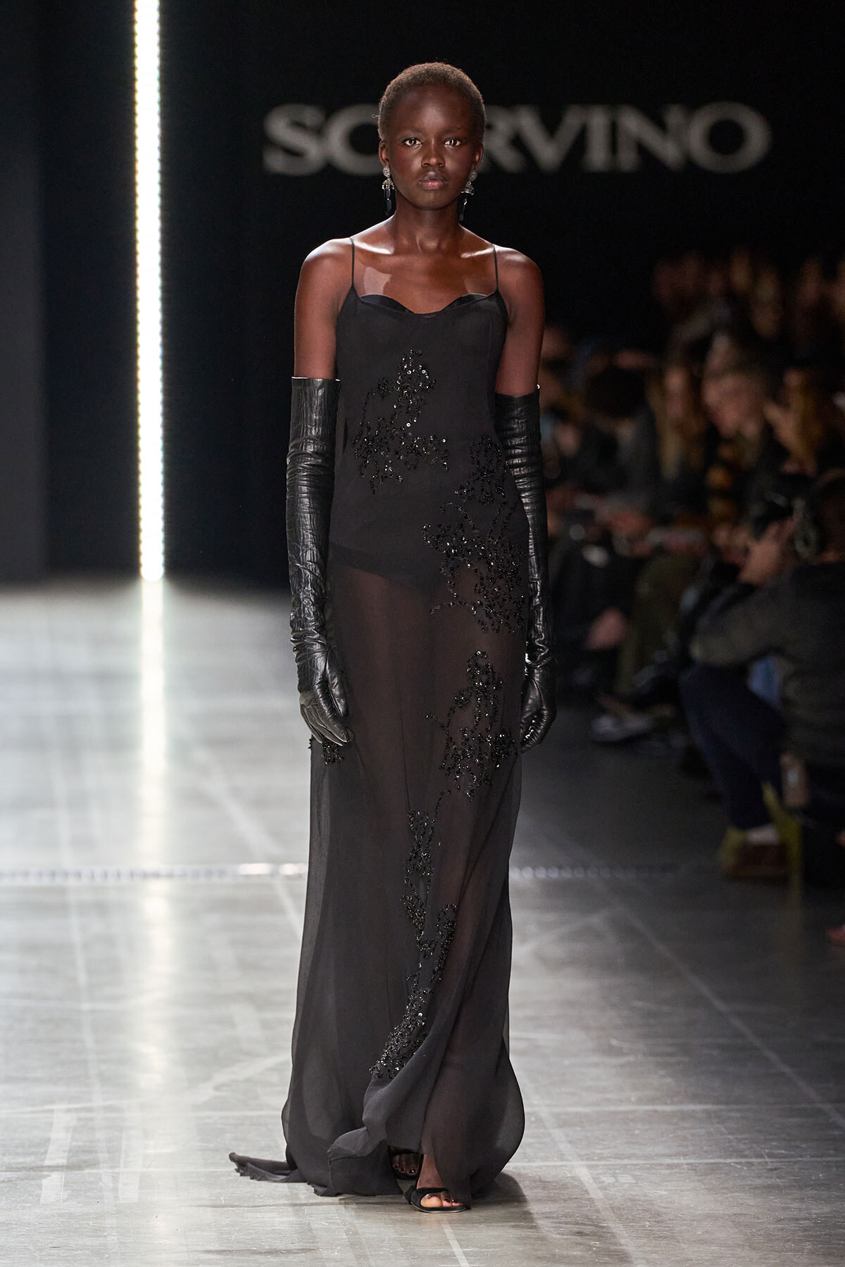 Model walks the runway in a black sheer evening gown with bead embroidery and long black gloves, against a fashion show backdrop.