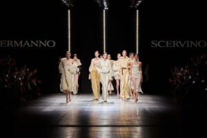 Group of models walk a fashion runway in cream and beige outfits as an audience watches from the sides; vertical lights glow in a dark backdrop.