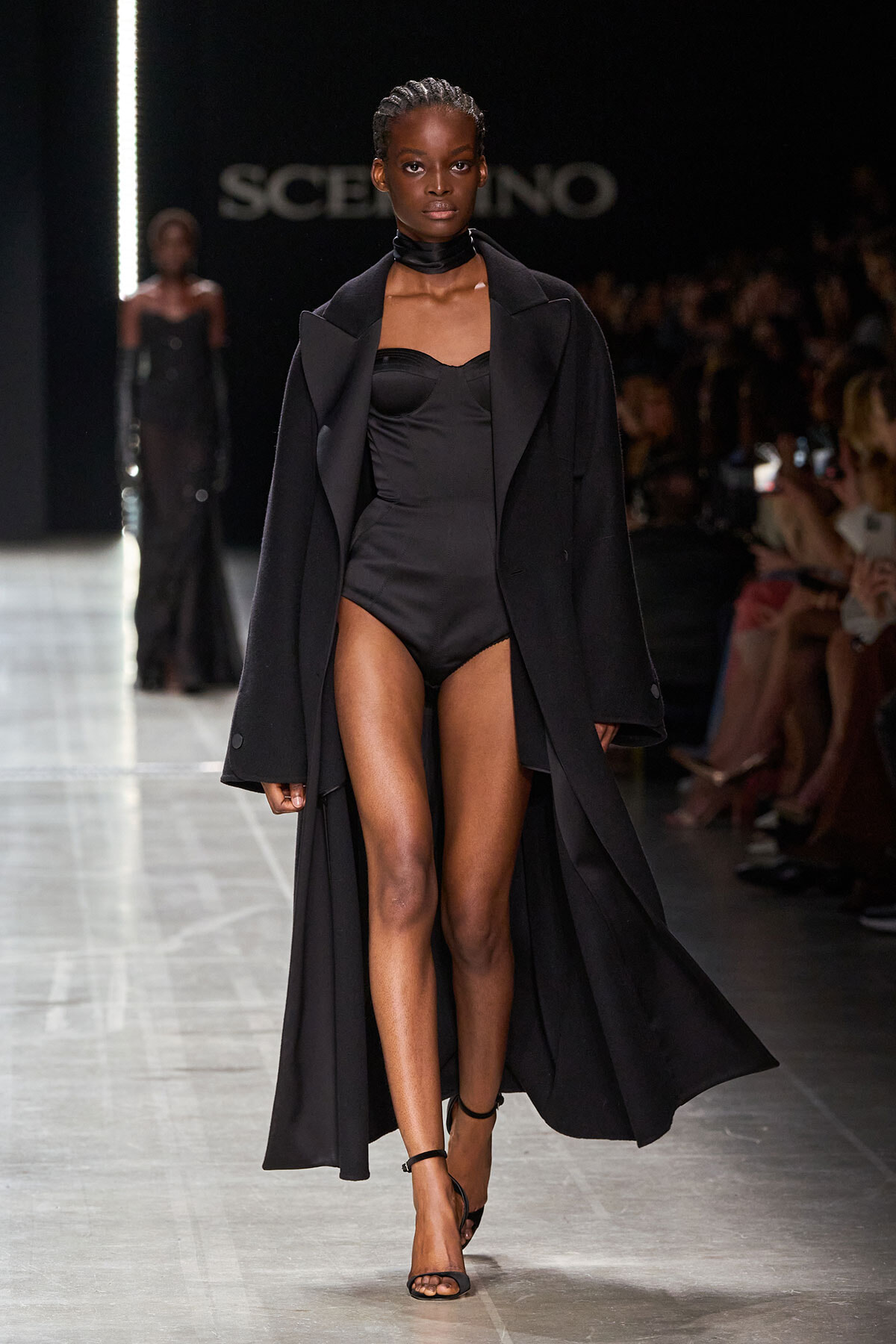 Model walks the runway in a black strapless bodysuit with a long black coat and black sandals, audience visible in the background.