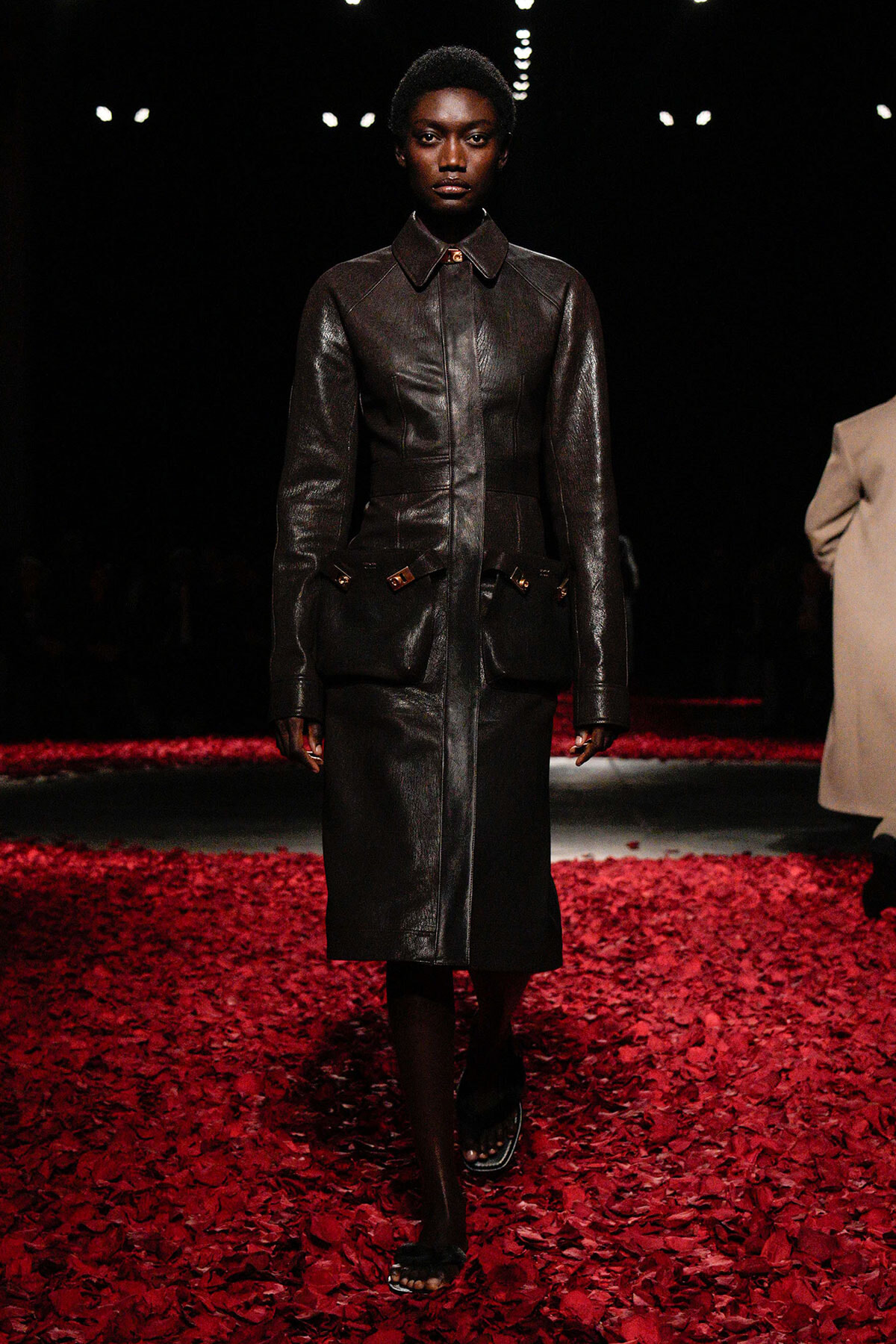 Model wearing a dark brown leather trench with oversized front pockets walks down a red petal-covered runway on a dimly lit catwalk.