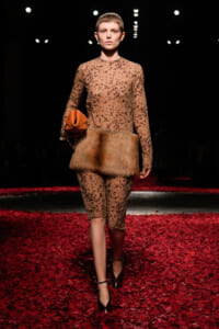Model walks the runway in a beige leopard-print sheer dress with a thick fur waist belt, carrying an orange clutch on a red petal-covered catwalk.