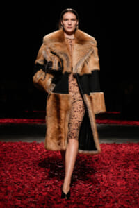 Model on a runway wearing a tan fur coat over a leopard-print dress; black paneling and red petal-covered floor beneath.