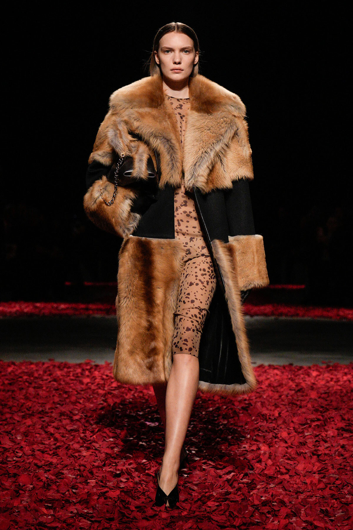 Model on a runway wearing a tan fur coat over a leopard-print dress; black paneling and red petal-covered floor beneath.