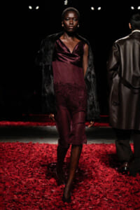 Model walking a fashion show runway in a burgundy satin dress with lace hem and a black faux-fur coat, surrounded by red petals on the floor.