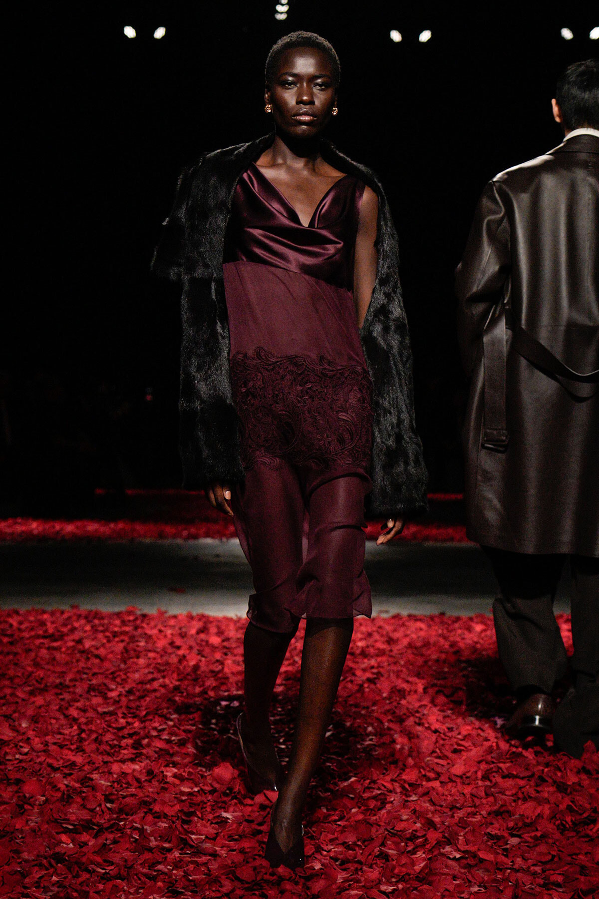 Model walking a fashion show runway in a burgundy satin dress with lace hem and a black faux-fur coat, surrounded by red petals on the floor.