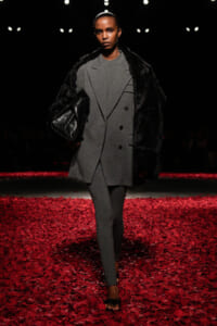 Model walking a runway in a gray, double‑breasted outfit with a black fur wrap and a large black leather bag; red rose petals cover the floor.