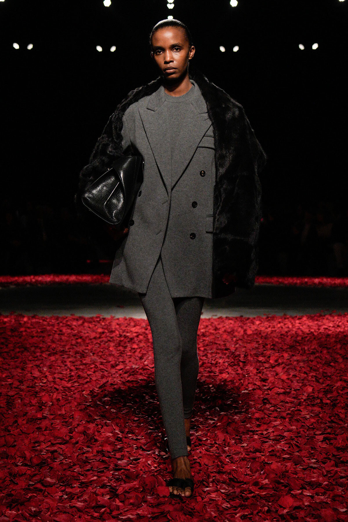Model walking a runway in a gray, double‑breasted outfit with a black fur wrap and a large black leather bag; red rose petals cover the floor.