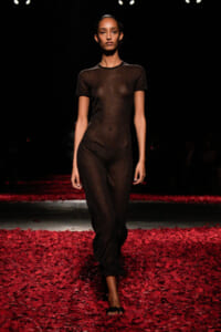 Model walking a runway in a sheer black bodysuit on a floor covered with red rose petals.
