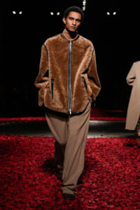 Male model on a runway wearing a brown plush zip-front jacket over a tan turtleneck and wide beige trousers, walking on a red petal-covered catwalk.
