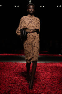 Model walking a fashion show in a tan leopard-print midi dress with a layered top, cinched waist, and a black clutch on a red-petaled runway.
