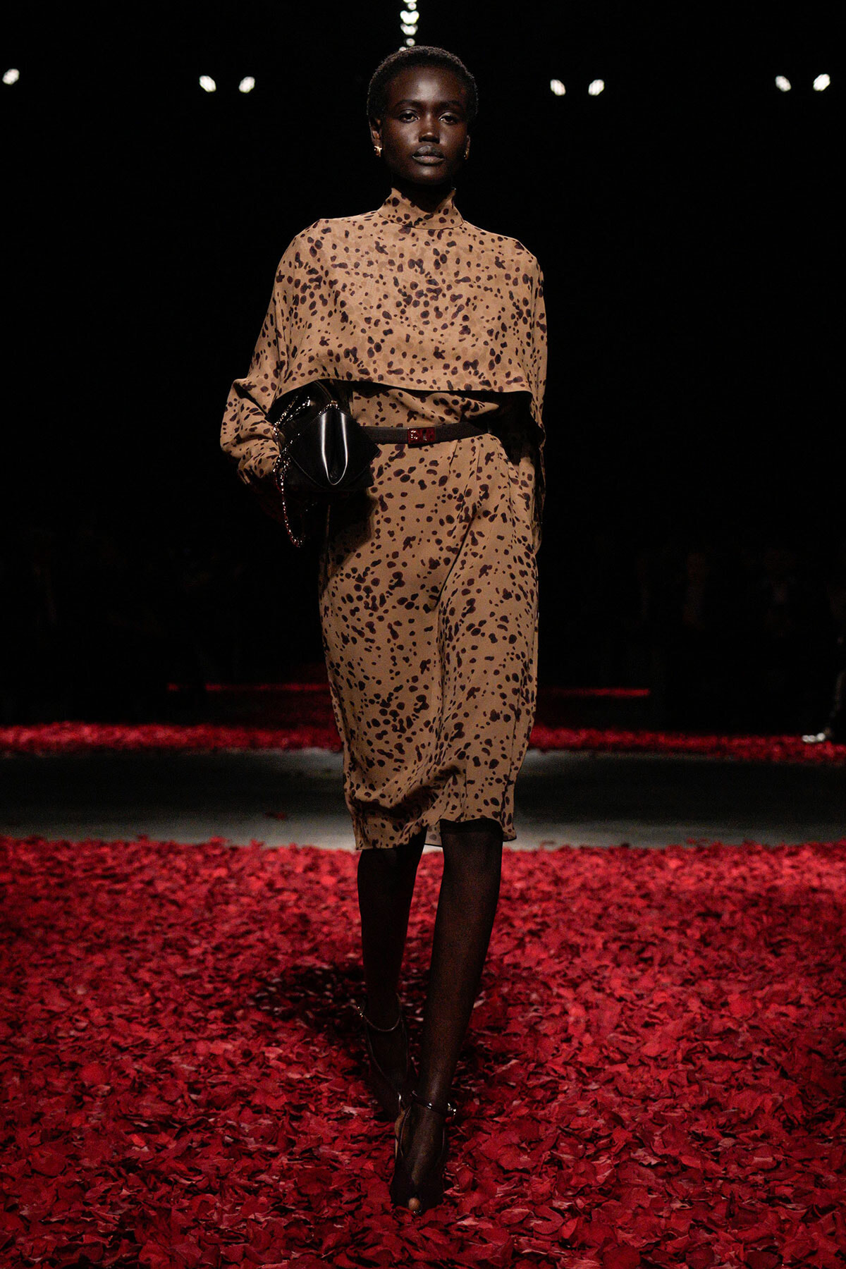 Model walking a fashion show in a tan leopard-print midi dress with a layered top, cinched waist, and a black clutch on a red-petaled runway.