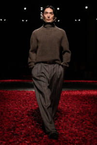 Male model walks a runway in a brown knit turtleneck and gray wide-leg trousers, hands in pockets, on a red-petaled catwalk with dark background.