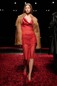 Model walks the runway in a red satin dress with lace detail, paired with a tan fur coat amidst red petal the runway.