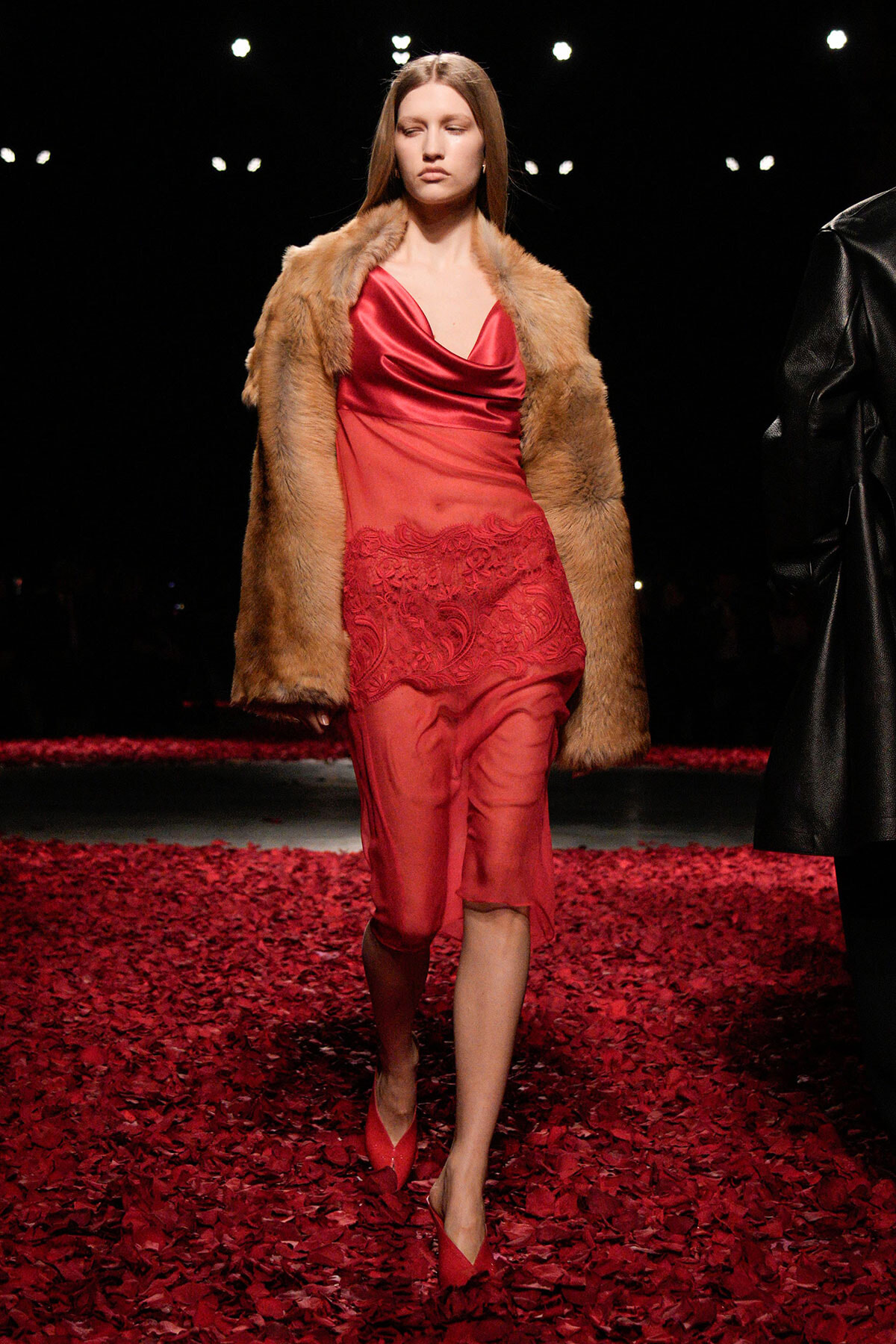 Model walks the runway in a red satin dress with lace detail, paired with a tan fur coat amidst red petal the runway.