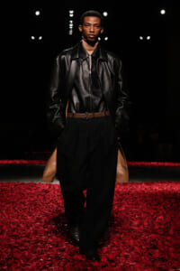 Male model on a runway wearing a black leather jacket, black pants with tan side panels, and a brown belt; red petals cover the catwalk.