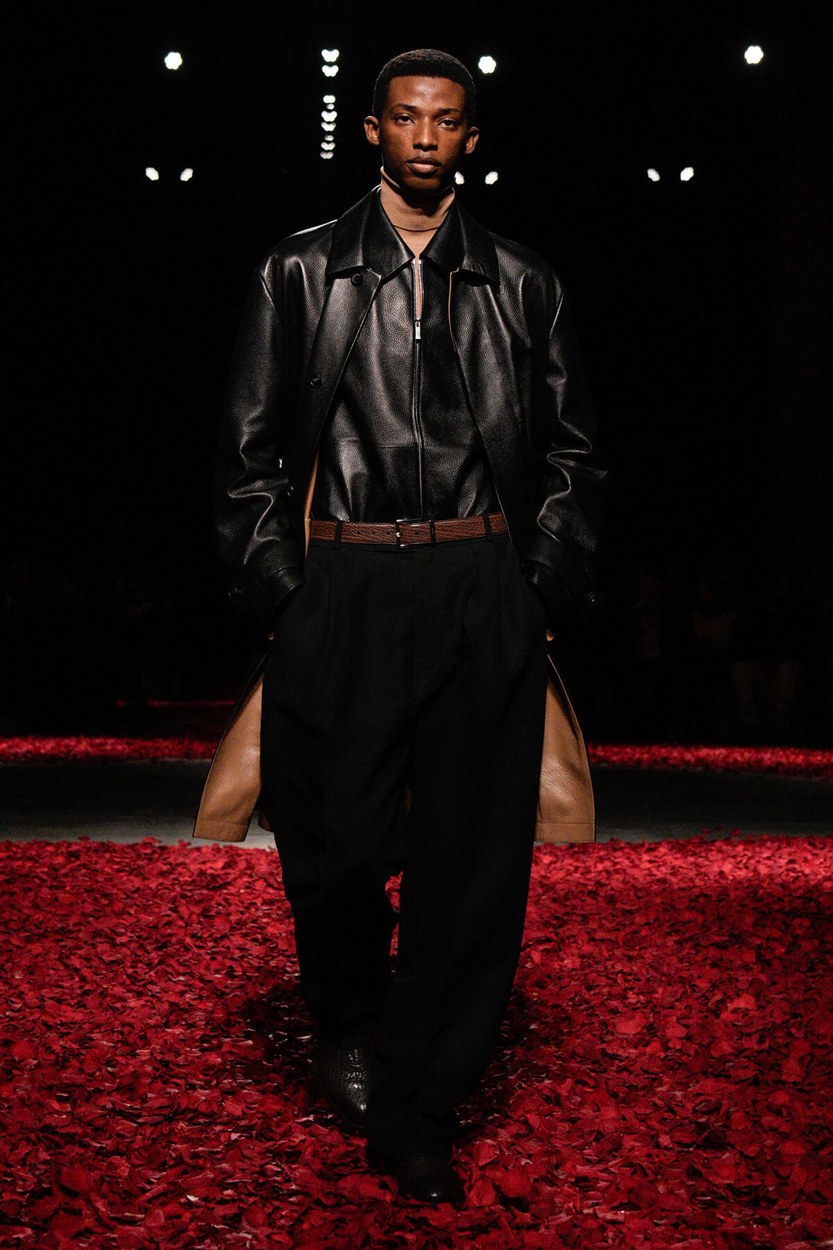 Male model on a runway wearing a black leather jacket, black pants with tan side panels, and a brown belt; red petals cover the catwalk.