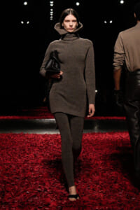 Runway model walks in a taupe knit turtleneck dress with matching leggings, carrying a black clutch on a red-petaled runway.