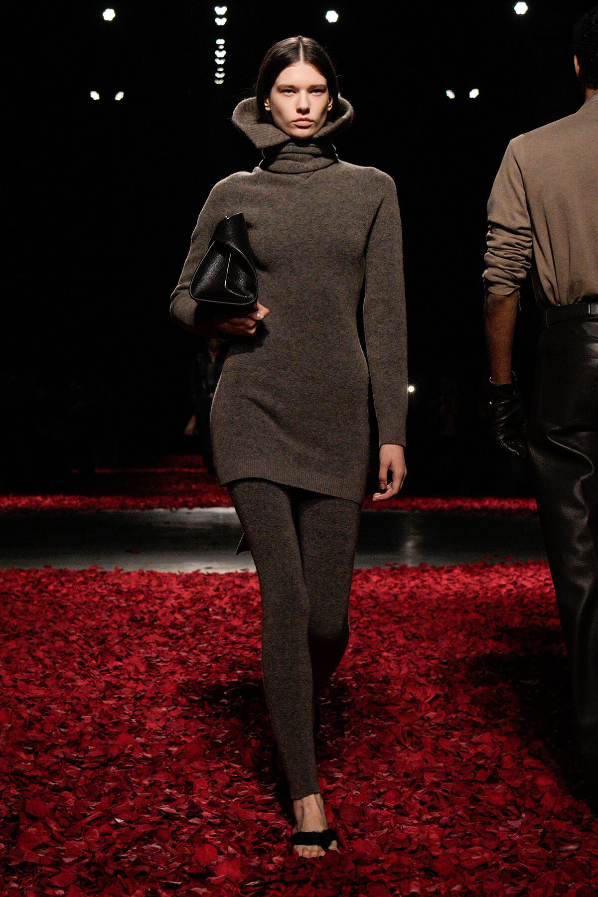 Runway model walks in a taupe knit turtleneck dress with matching leggings, carrying a black clutch on a red-petaled runway.