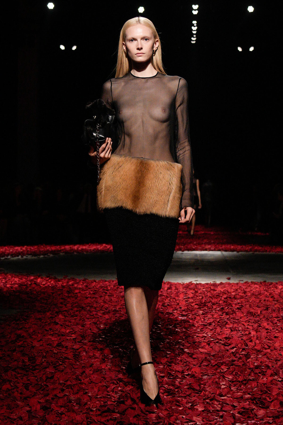 Female model on a dim runway wearing a sheer black top and a tan fur belt over a black knee-length skirt, with black heels, walking on red-petaled flooring.