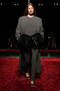 Model on a fashion runway in a gray oversized coat and matching outfit, holding two large black handbags at her waist amid a red petal floor.