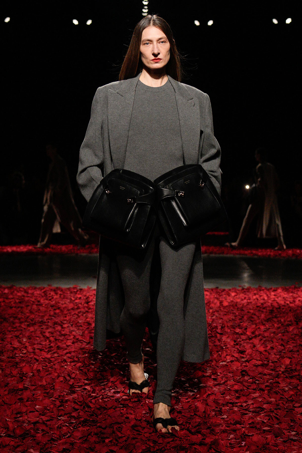 Model on a fashion runway in a gray oversized coat and matching outfit, holding two large black handbags at her waist amid a red petal floor.