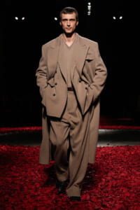 Male model walking a fashion runway in a tan suit with a long overcoat, on a red-petaled catwalk backdrop.